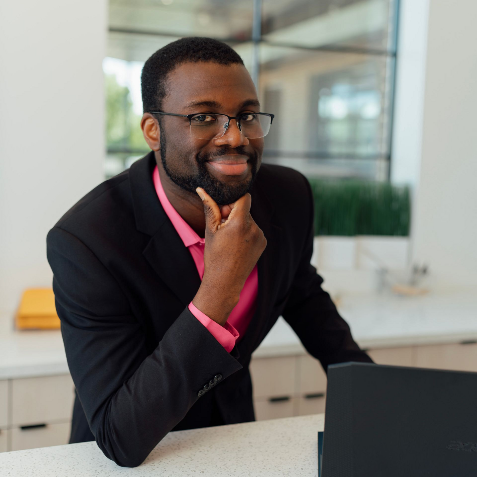 Man in black blazer and pink shirt, resting chin on hand, looking at camera with laptop in front.