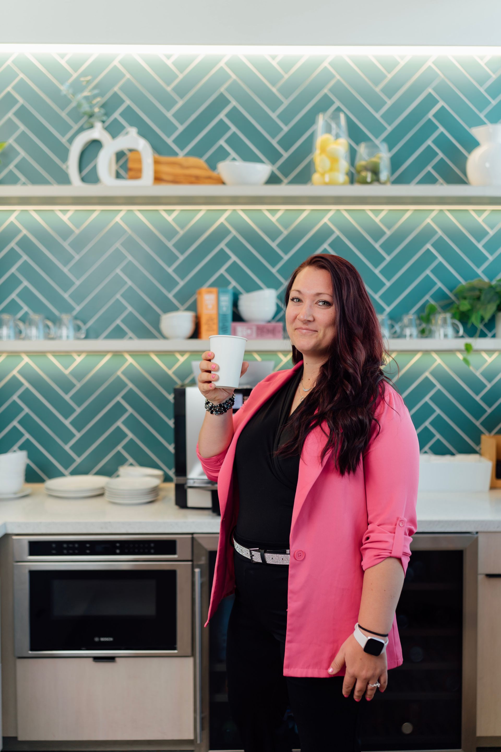 Woman in pink blazer holding coffee, standing in office kitchen with teal backsplash.