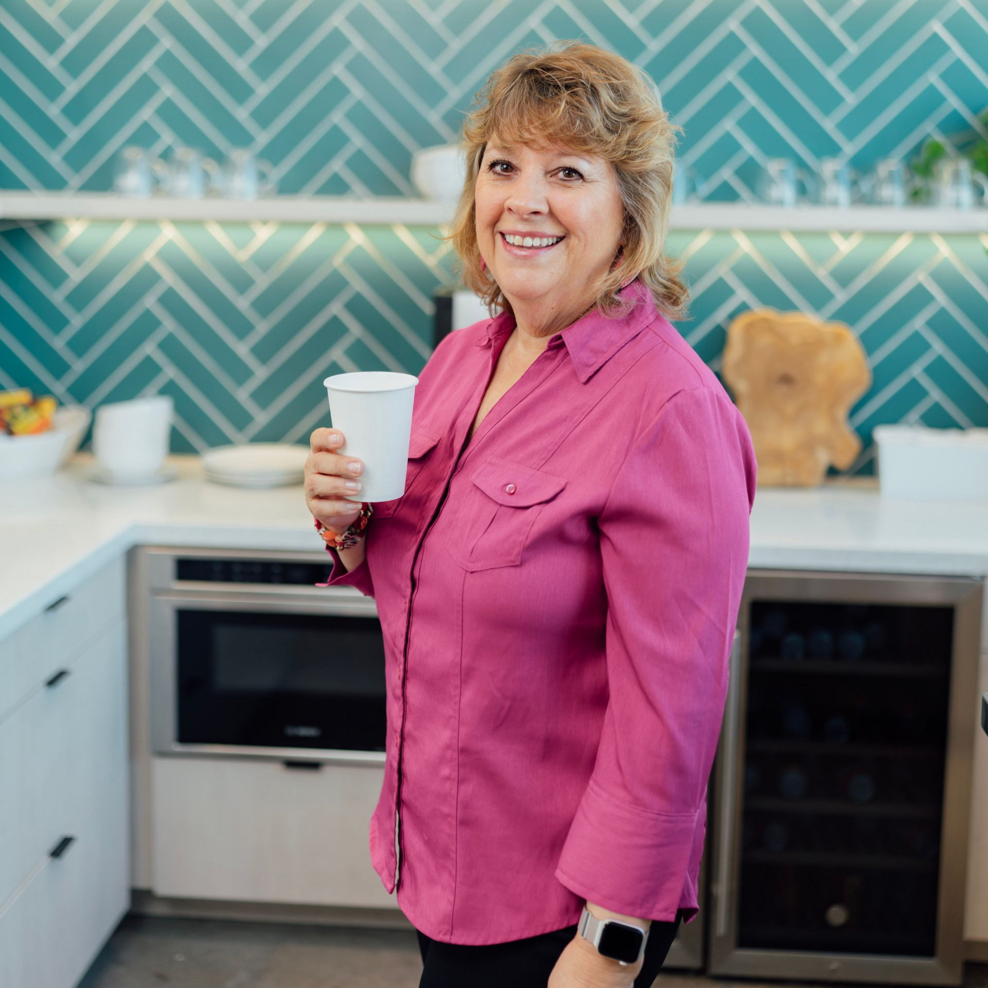 Woman in pink shirt holding a cup in a kitchen with turquoise tile backsplash.