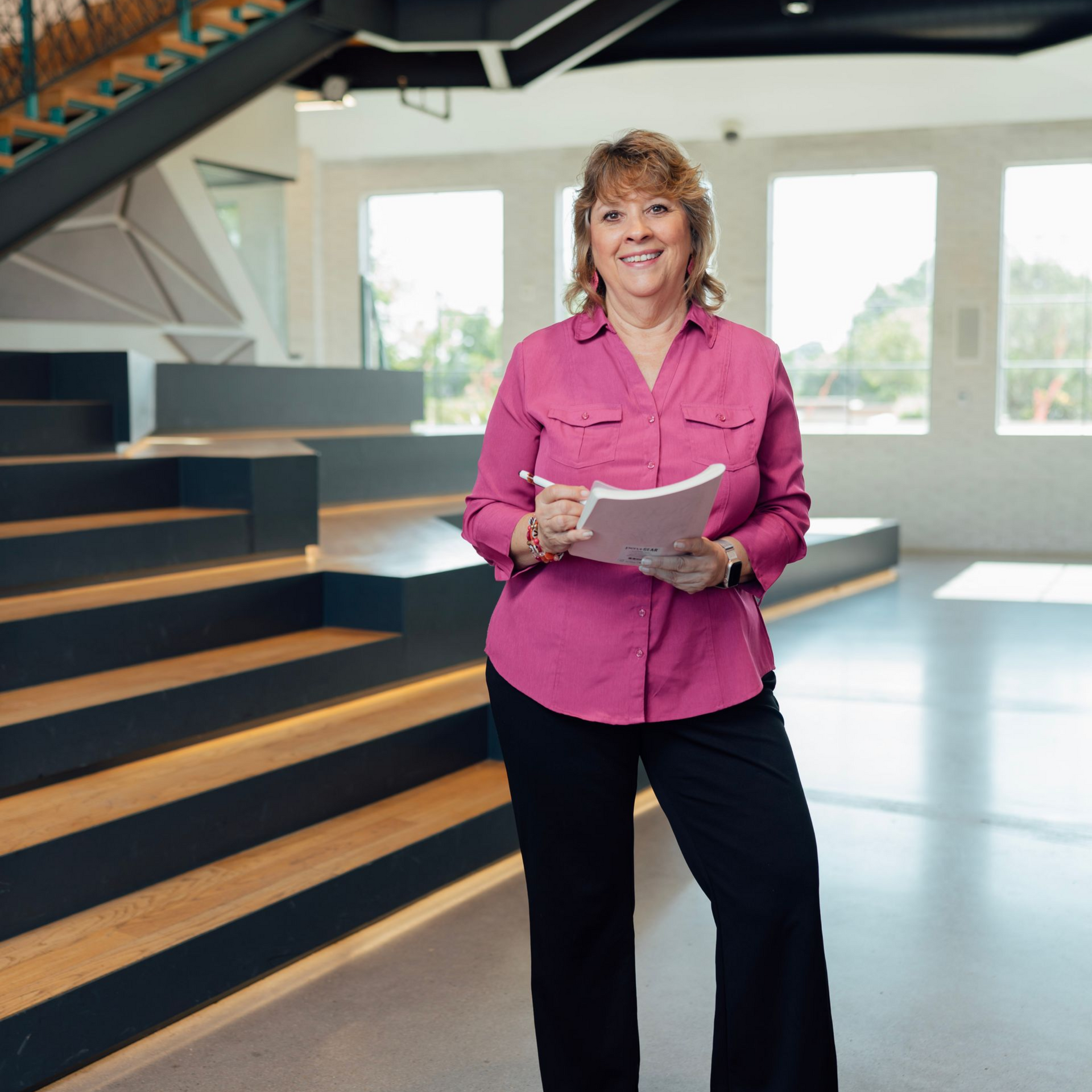 Woman in pink shirt and black pants holding a notepad and pen, smiling in a modern office.