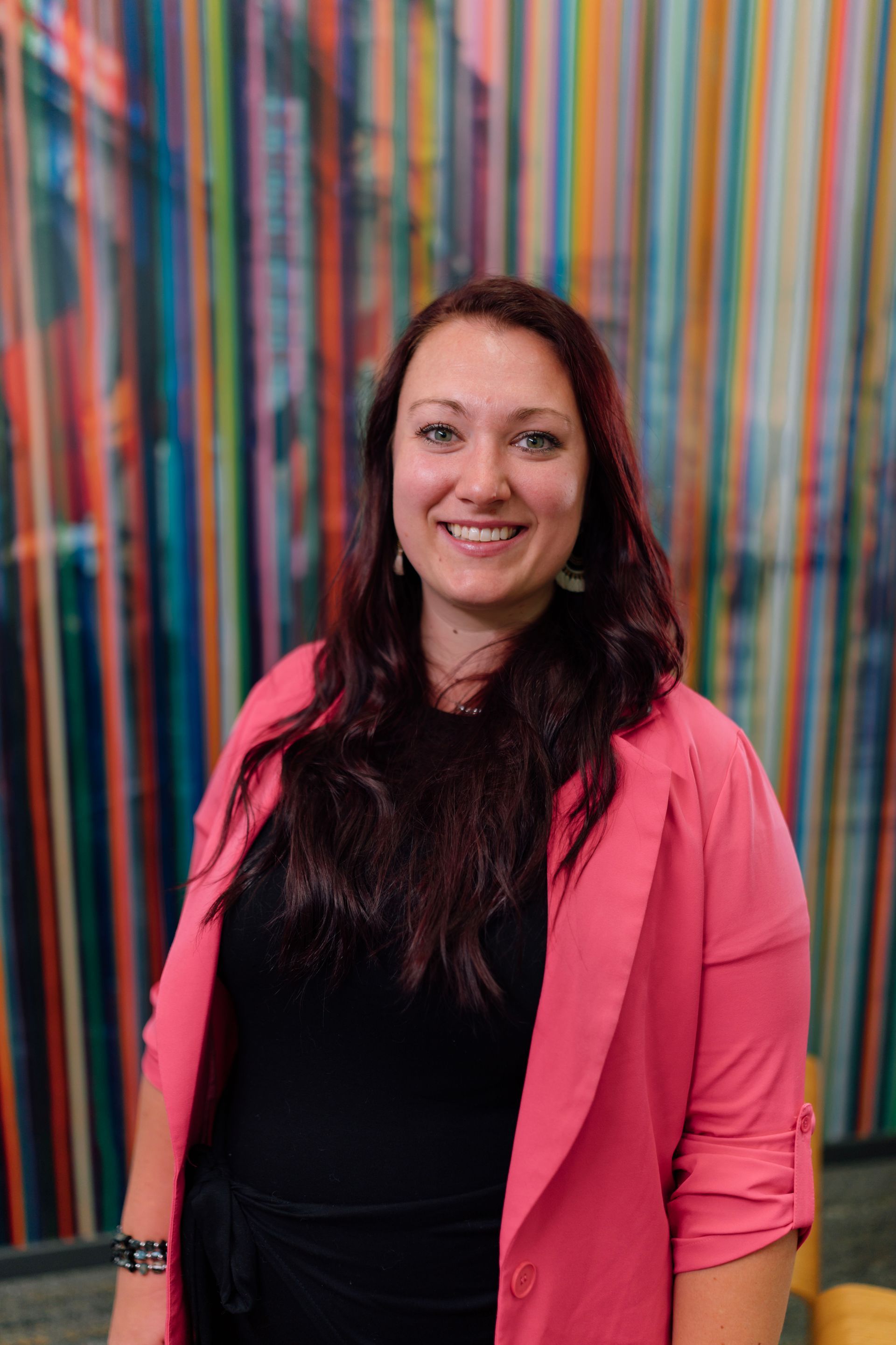 Woman with dark red hair, wearing a pink blazer and black top, smiles in front of colorful striped wall.