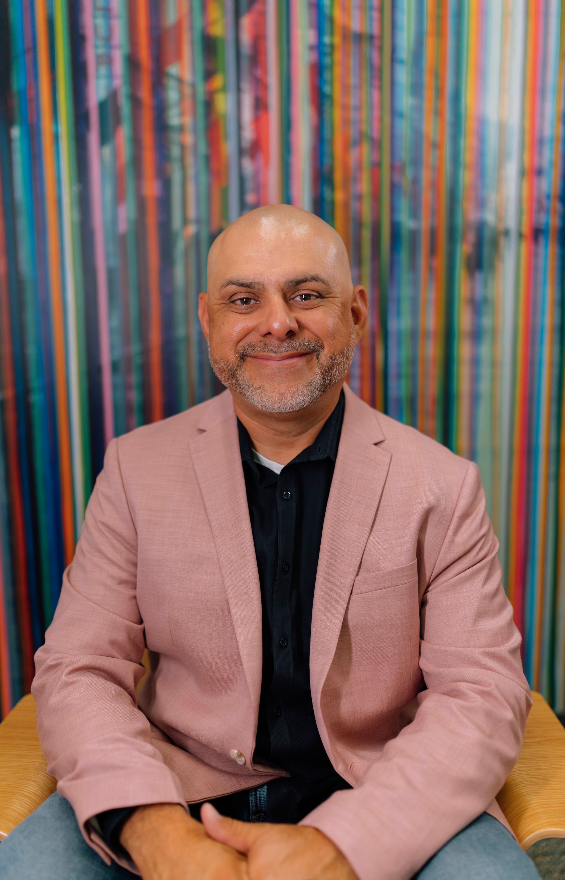 A man in a suit and pink shirt is standing with his arms crossed and smiling.