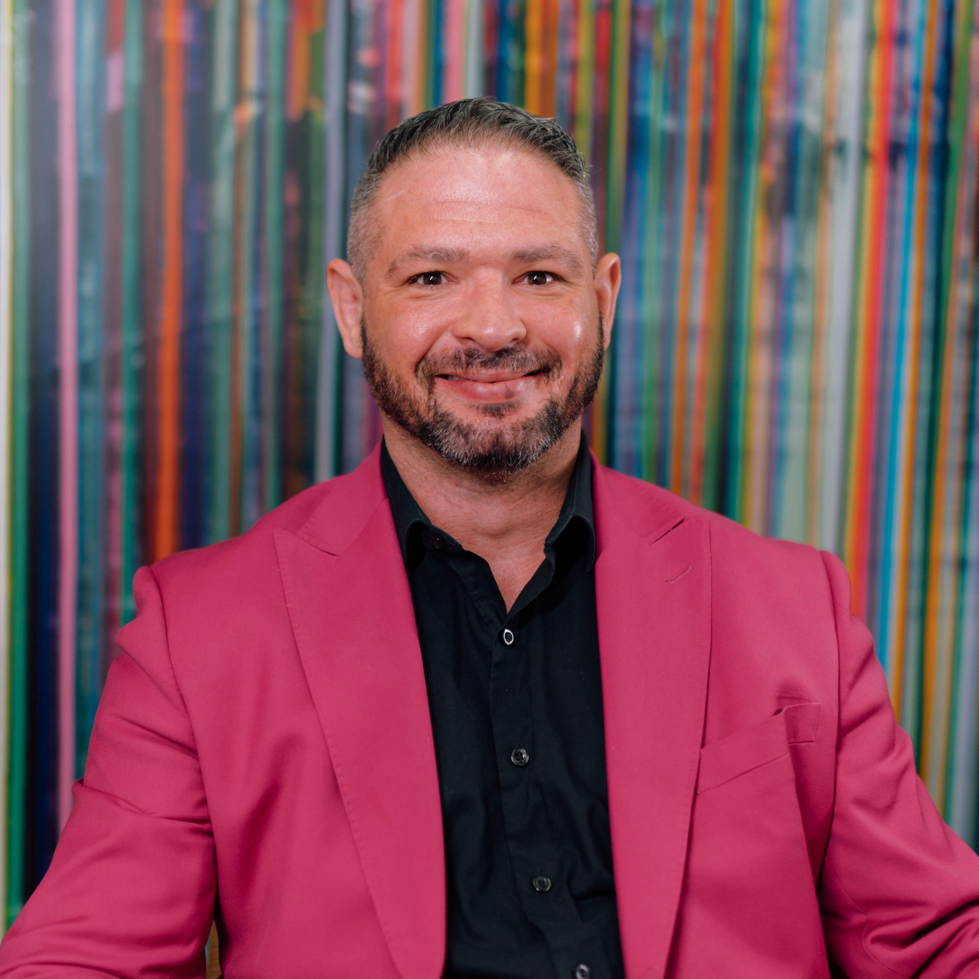 Man in pink suit smiles, black shirt, against a colorful striped background.