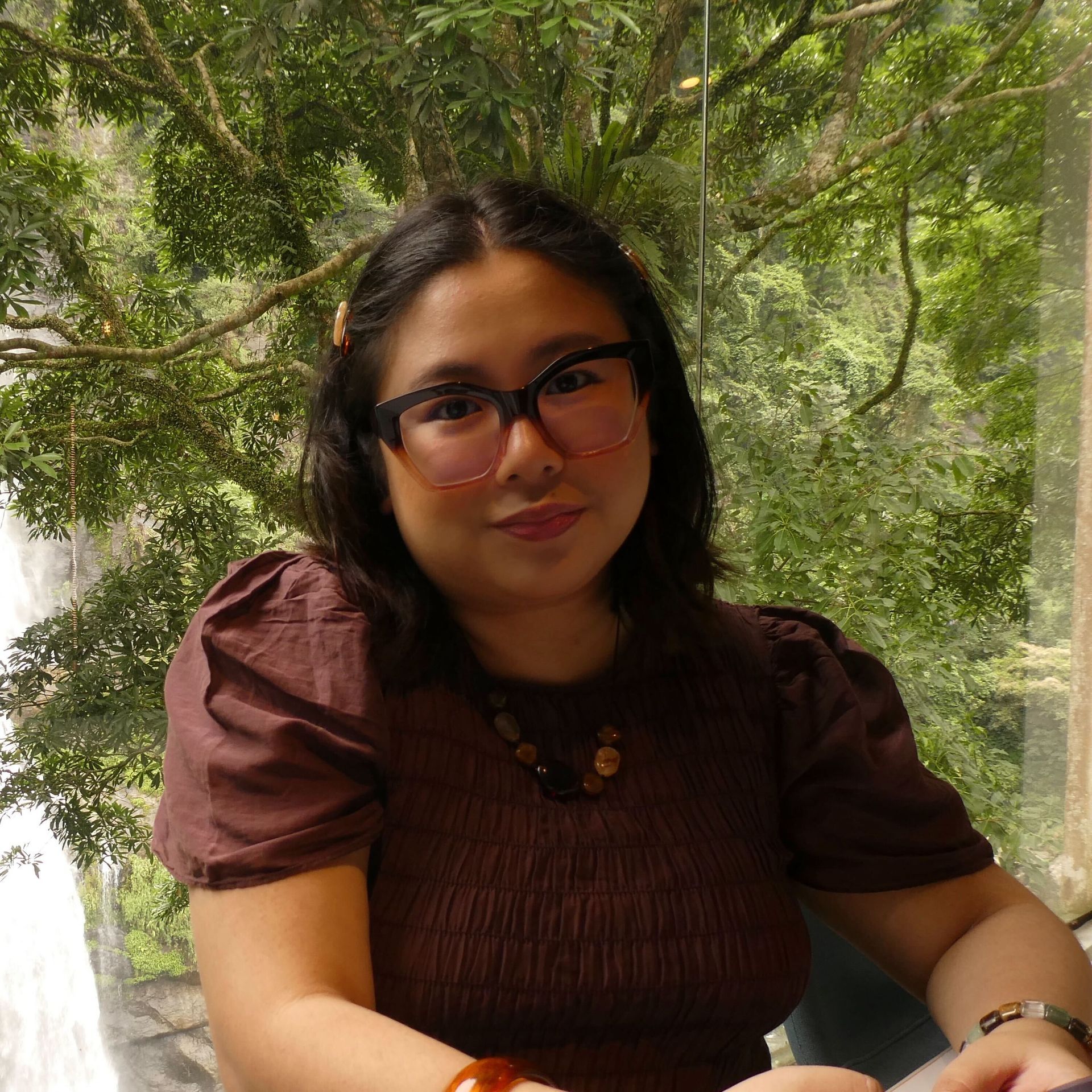 Woman with glasses and brown shirt smiles in front of a window with a waterfall view.