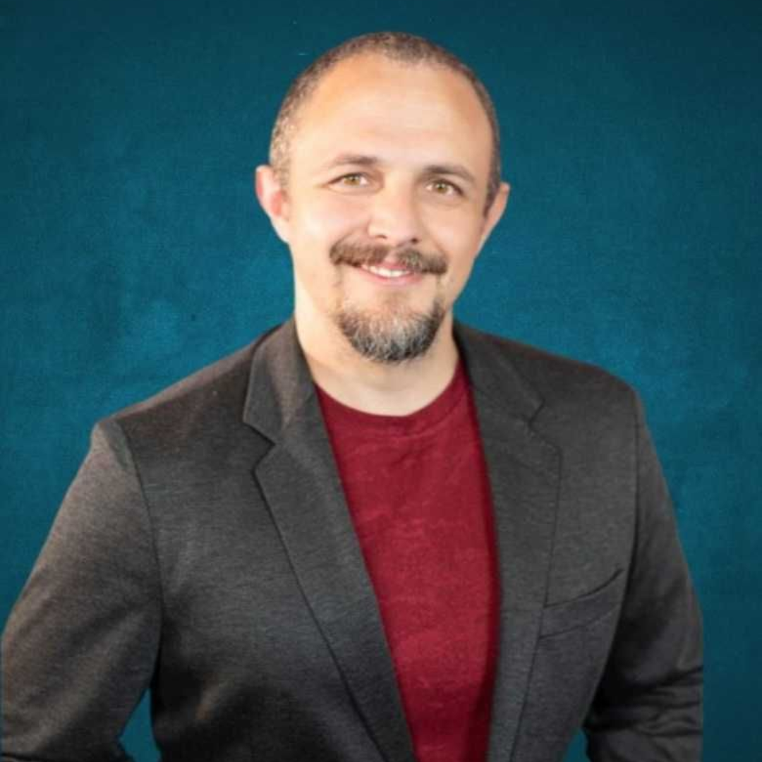 Man in gray blazer and red shirt smiling against a blue background.