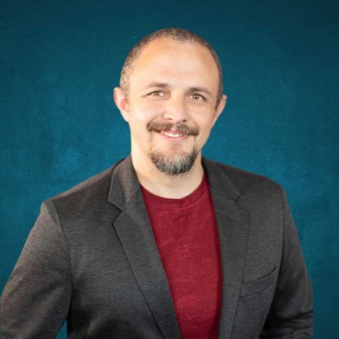 Man in red shirt and gray blazer smiles, holding jacket open against a teal backdrop.