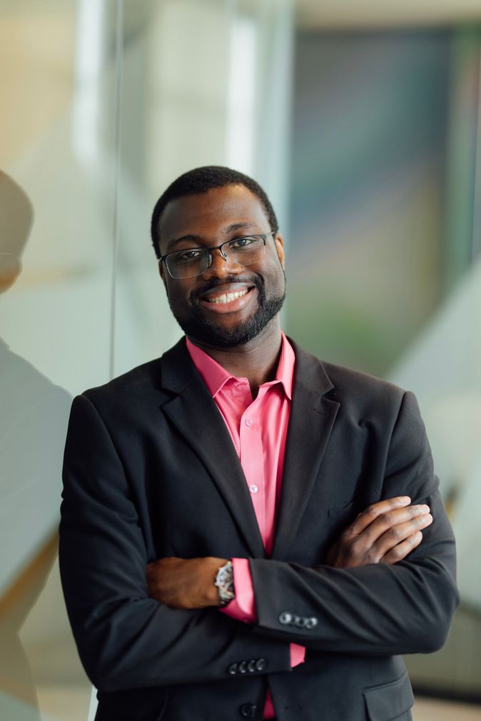 A man in a suit and pink shirt is standing with his arms crossed and smiling.