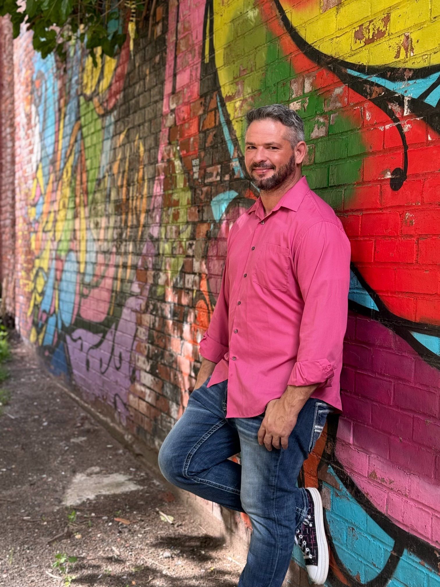 A man in a pink shirt is leaning against a colorful graffiti wall.