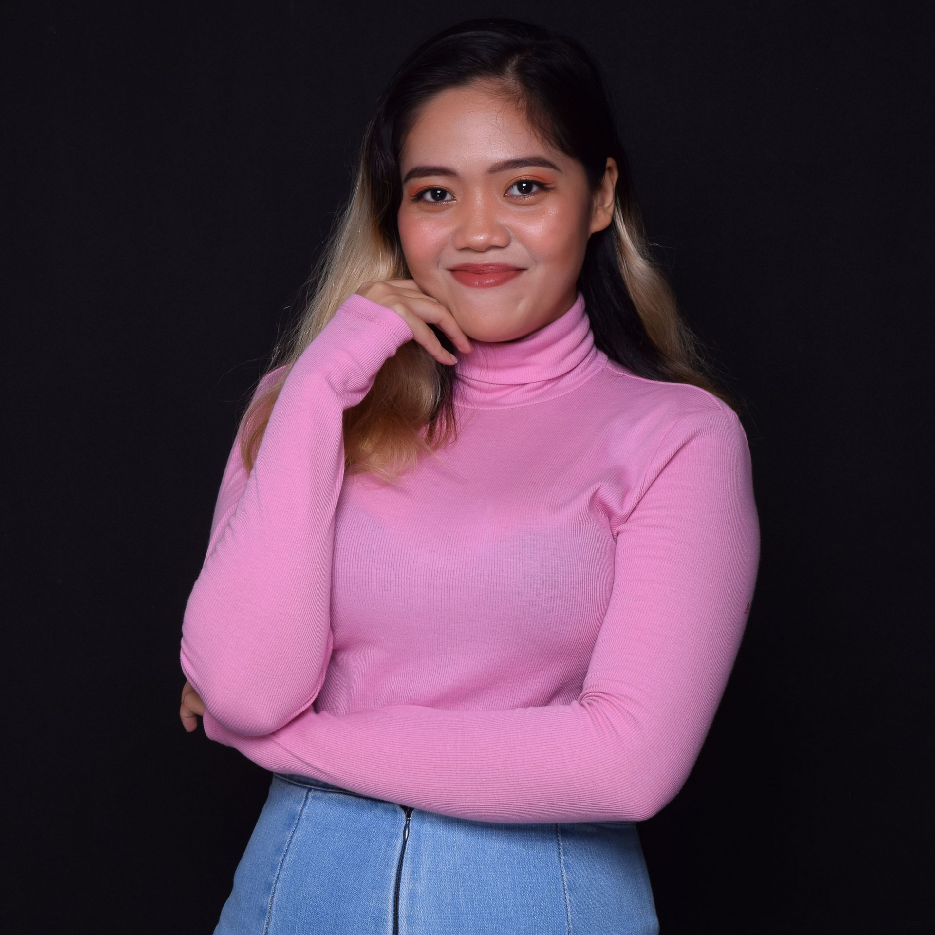 Woman in a pink turtleneck and denim skirt, hand on chin, smiles. Black background.