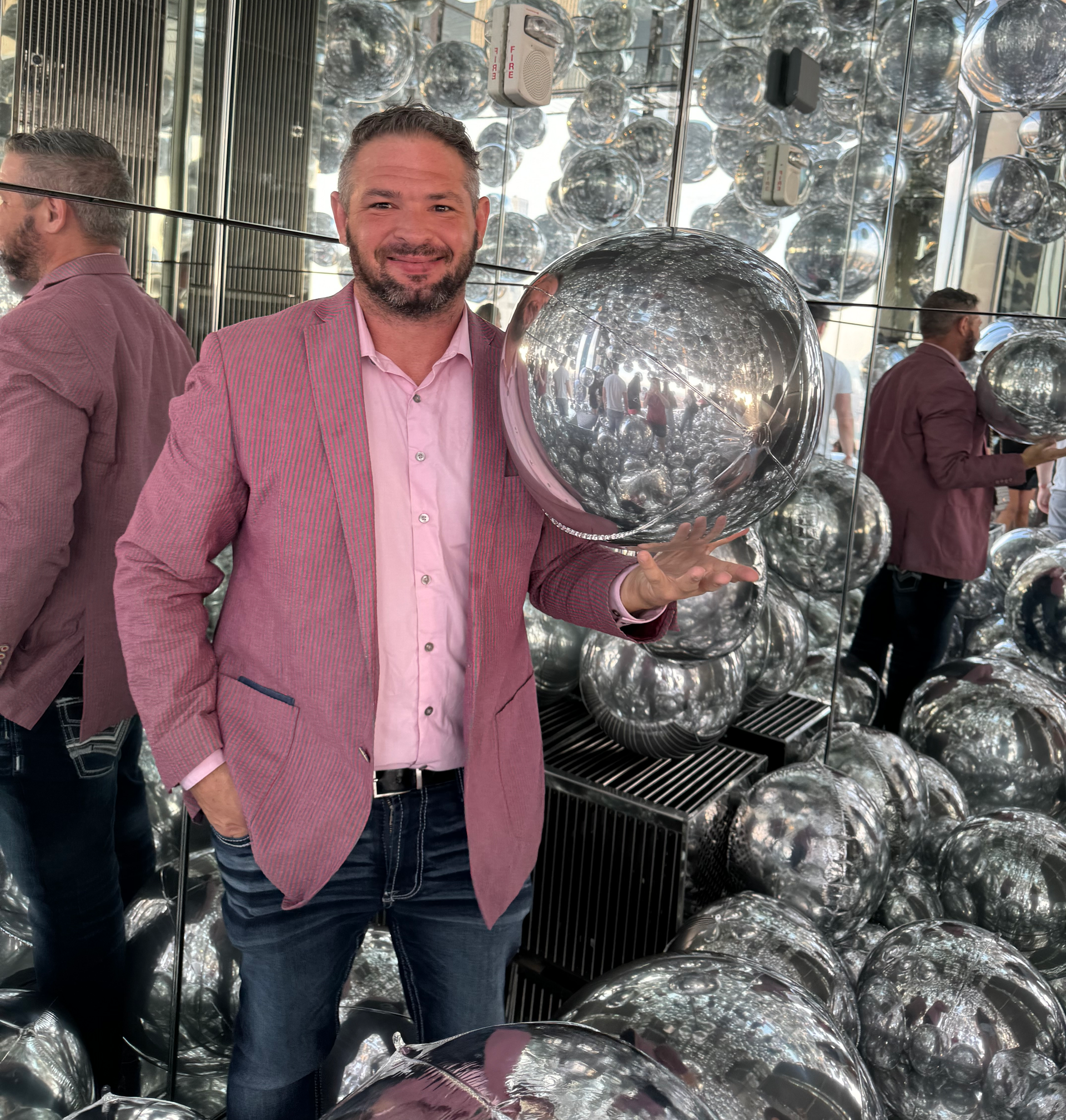 A man in a pink jacket is holding a silver balloon in front of a wall of mirrors.