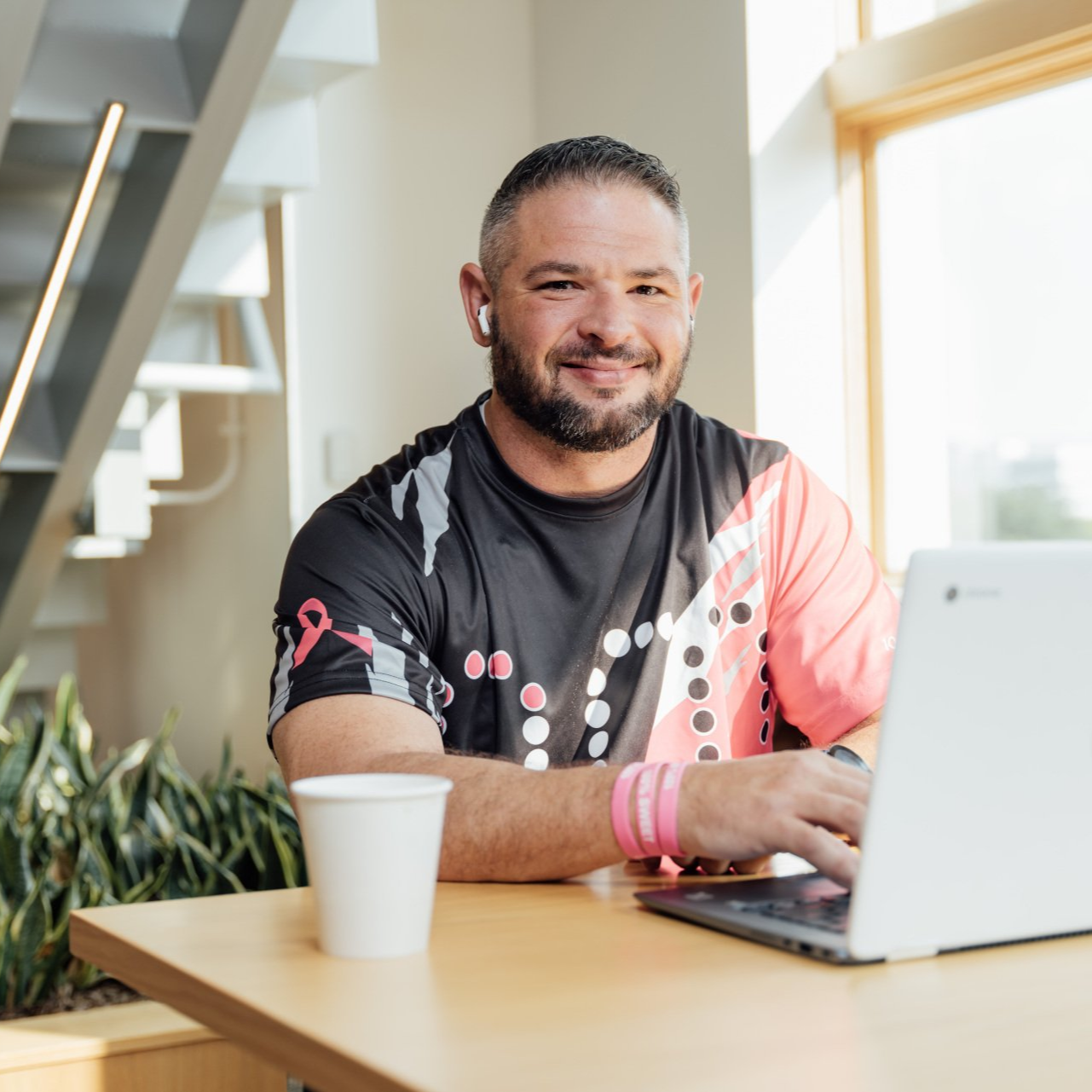 Man working on a laptop at a table, smiling. Indoors, next to a window and plants.