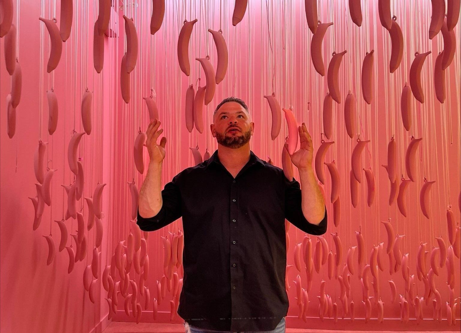 a man is standing in front of a pink wall with bananas hanging from the ceiling .