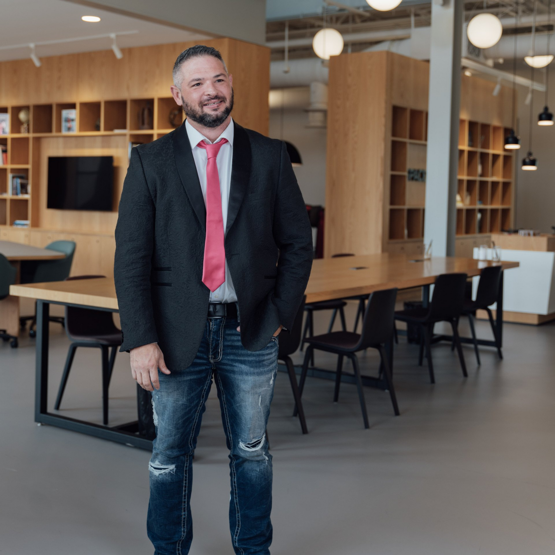 Man in blazer and jeans smiles in a modern office, with open shelving, tables, and round lights.