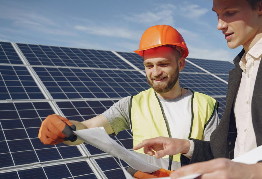 Two men reviewing blueprints in front of solar panels. One in a hard hat and vest, the other in a suit.