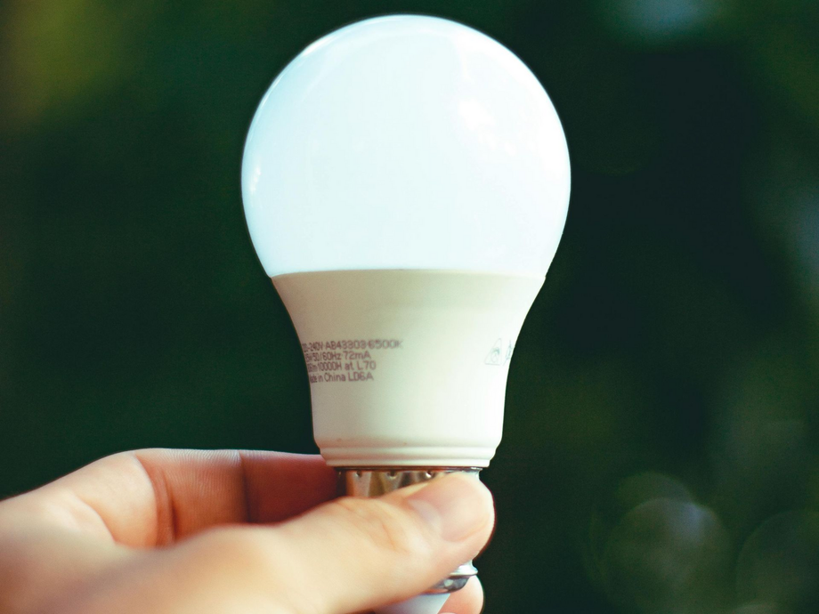 Hand holding a lit white LED lightbulb against a blurred green background.