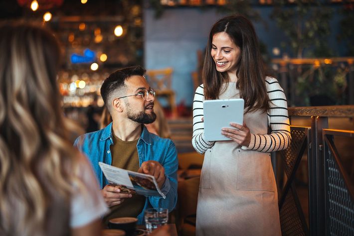 A woman is holding a tablet and talking to a man in a restaurant.