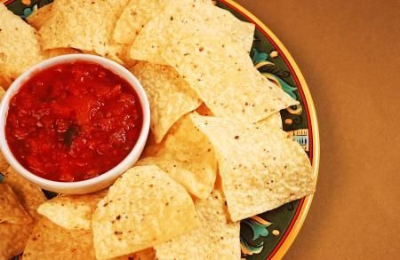 A bowl of red salsa sits on a decorative plate filled with light-colored tortilla chips on a brown surface.