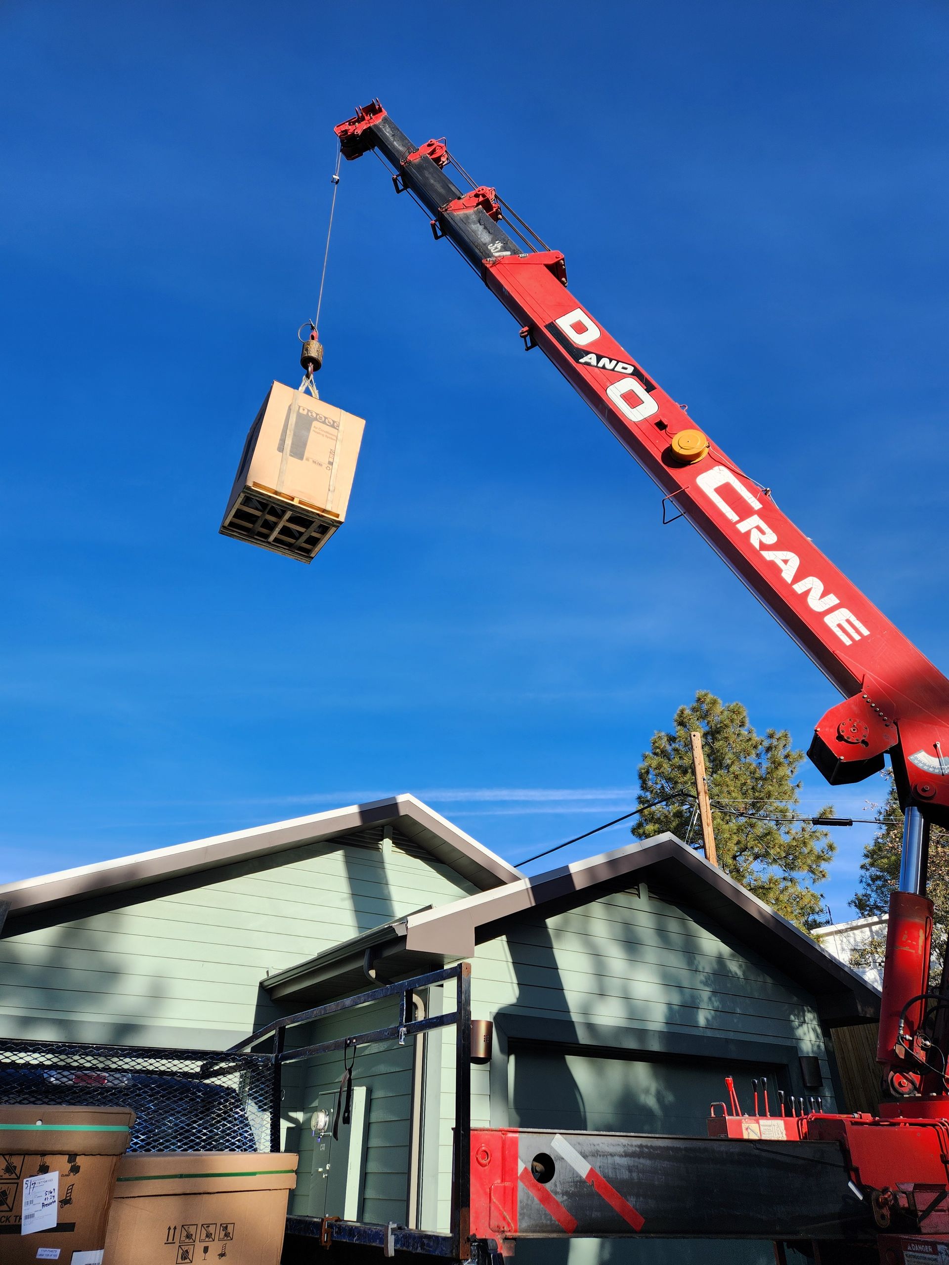 Red crane lifting a wooden crate against a blue sky, near a green house.