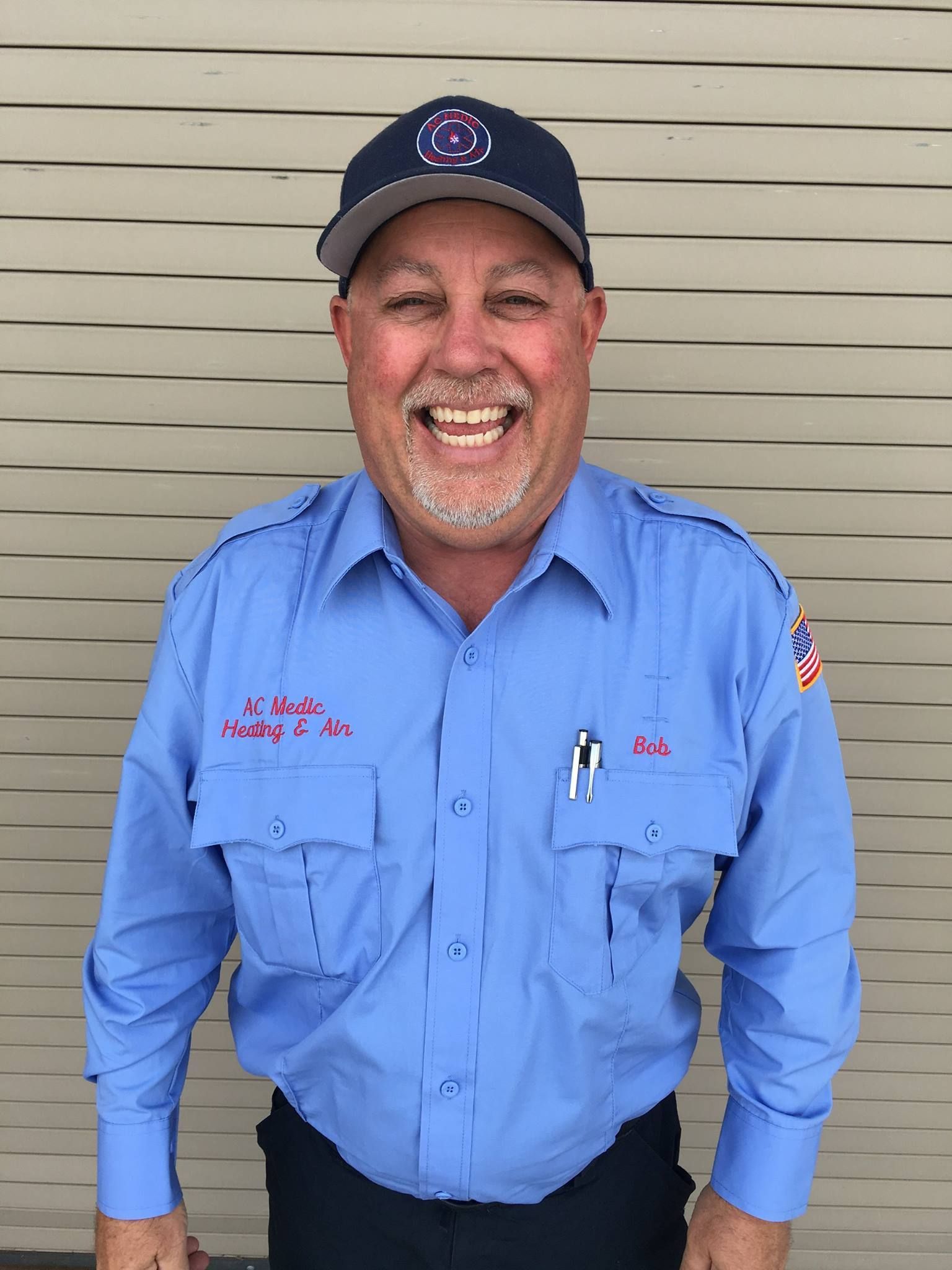 Man in blue uniform smiles, wearing a cap. He stands in front of a tan wall.