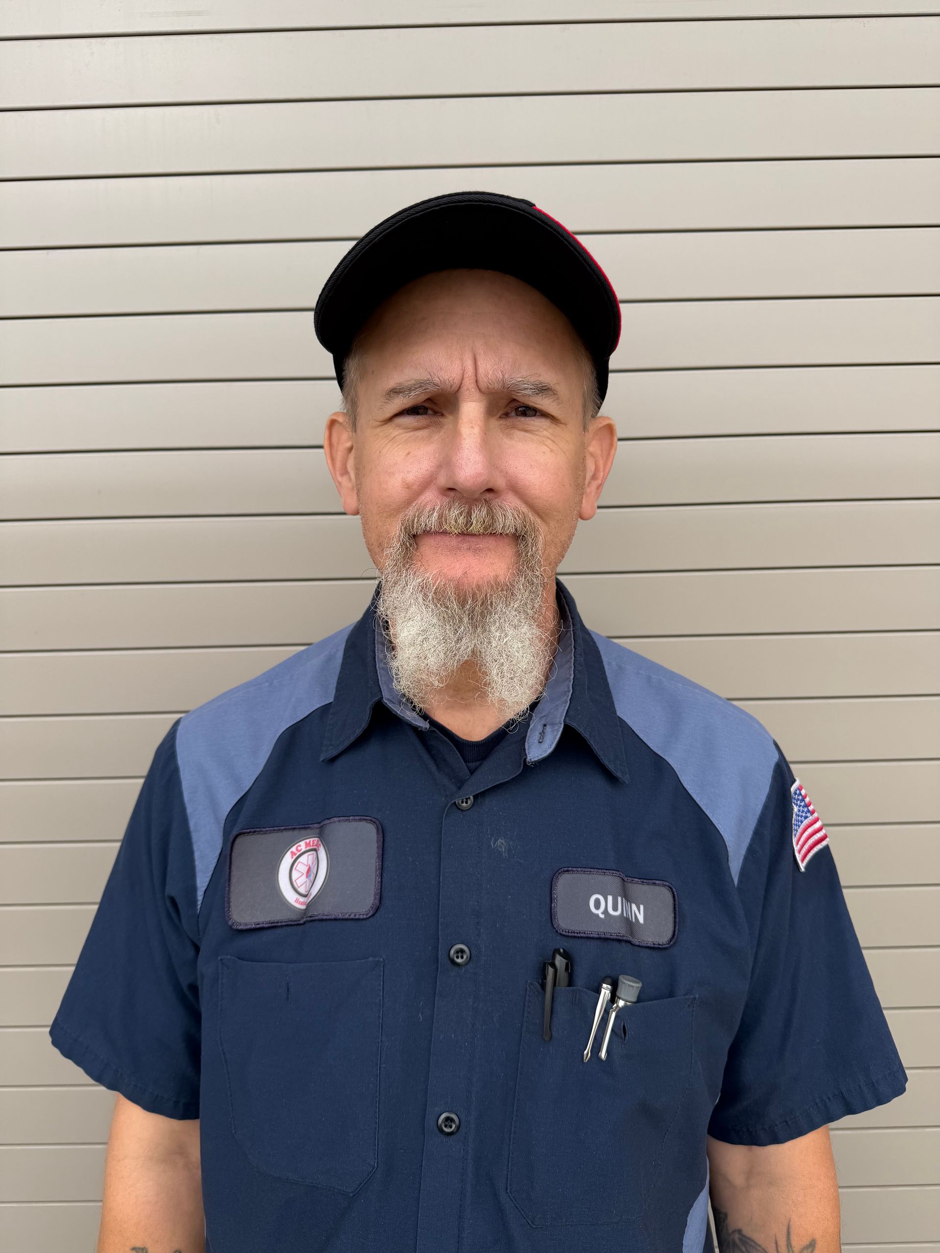 Man in mechanic uniform with a beard, wearing a hat, standing in front of a neutral background.