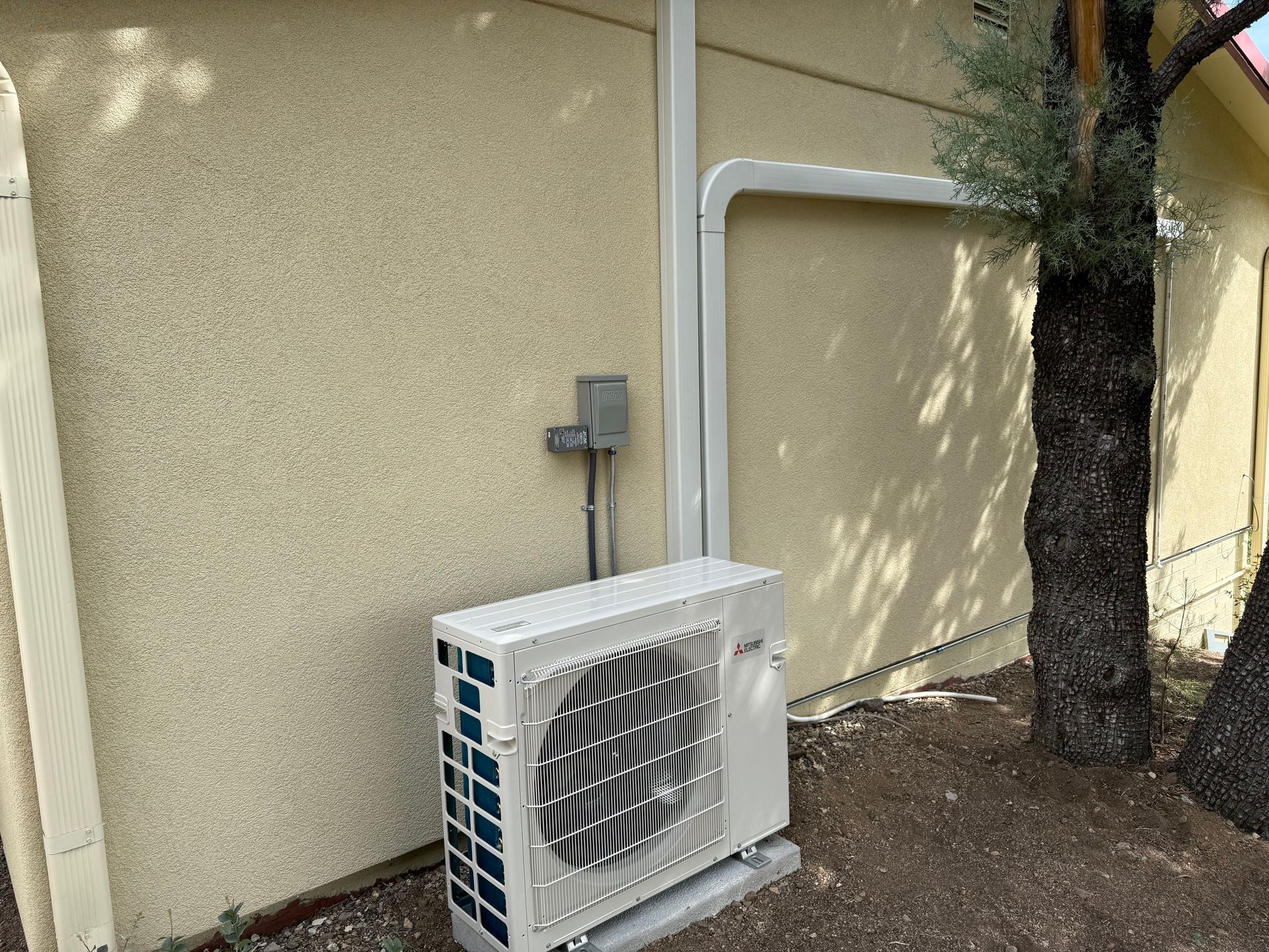 Outdoor air conditioning unit beside a stucco building, with electrical conduit and a tree nearby.