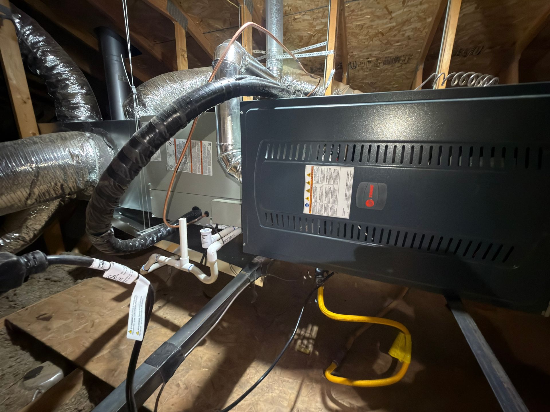 Furnace in an attic. Black appliance with ductwork and yellow extension cord on a wooden platform.