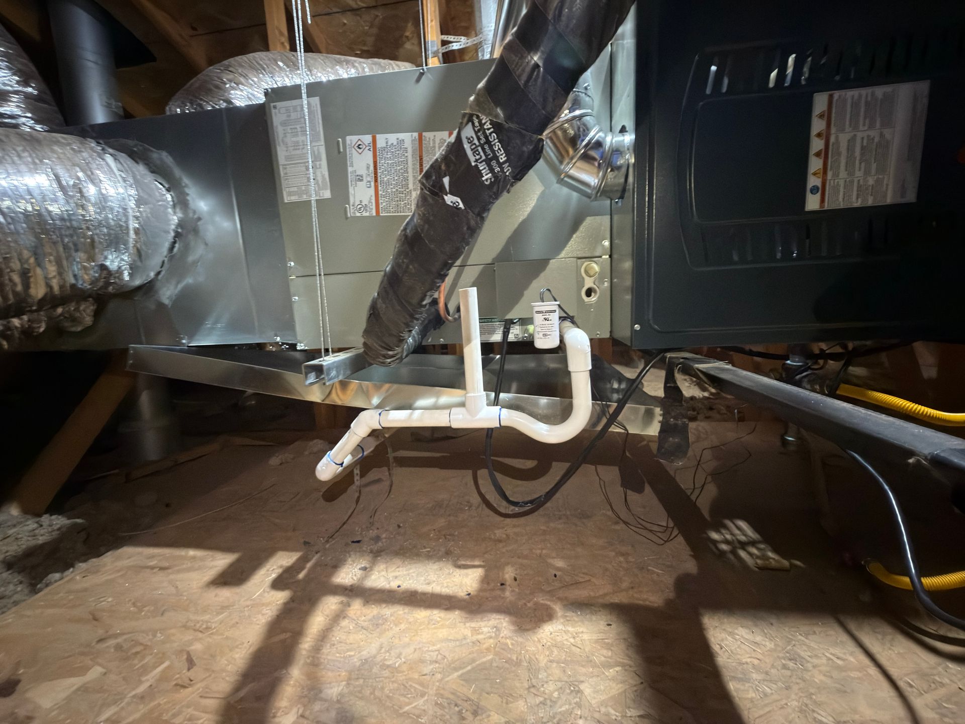 HVAC system in an attic. Ductwork, drain pipes, and electrical wiring visible. Wooden platform.