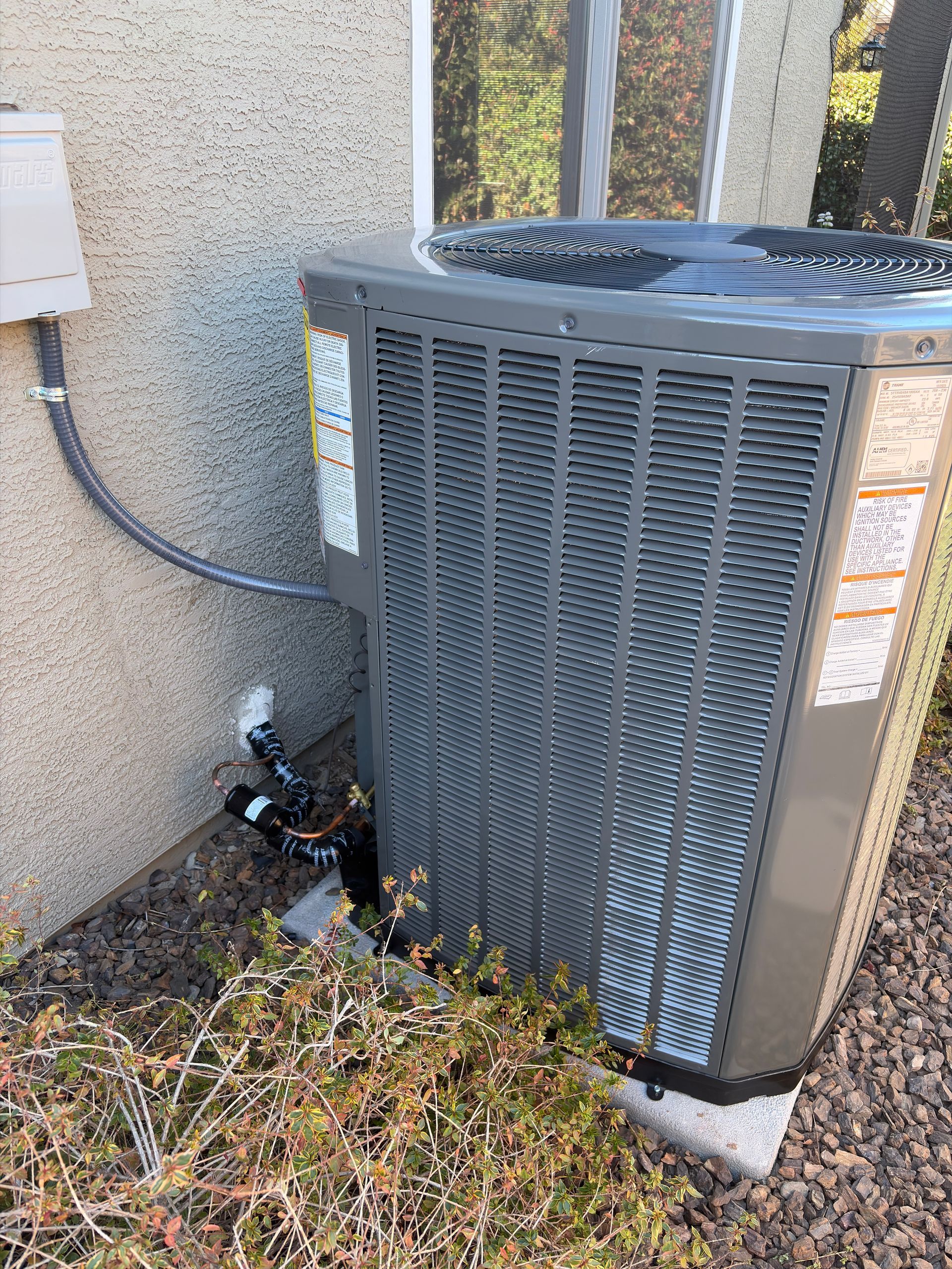Gray HVAC unit next to a stucco building with wiring and bushes in front.