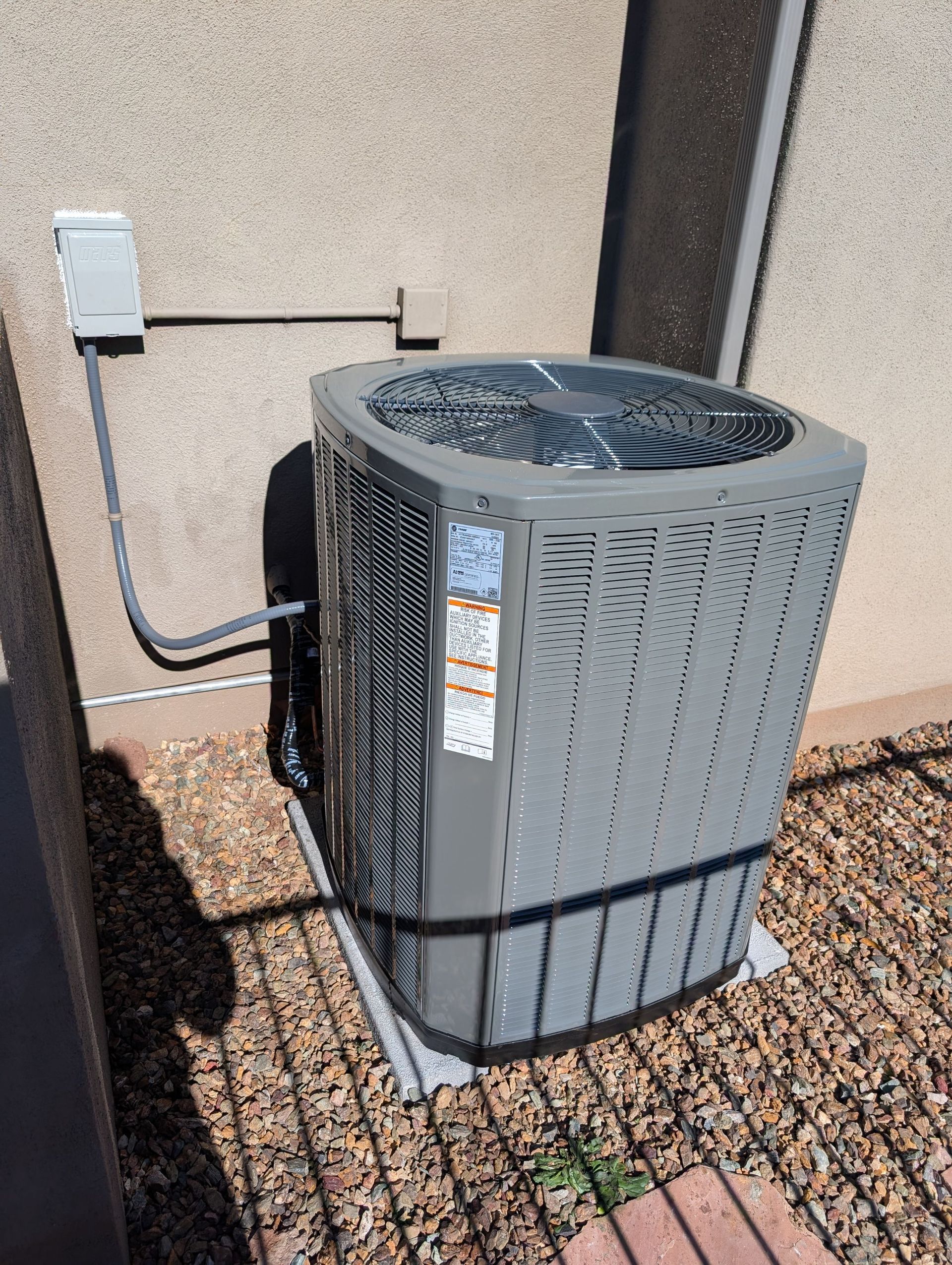 Air conditioner unit, gray, against a tan stucco wall.  A metal grid base is underneath.