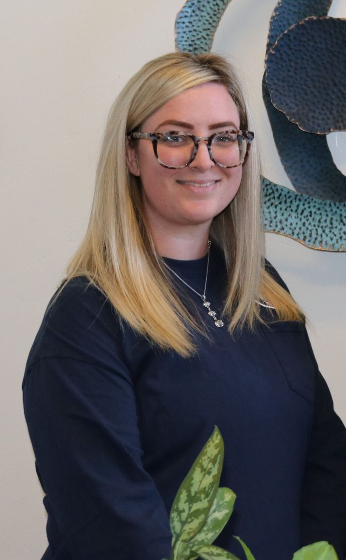 Woman with glasses smiles, wearing a navy sweater. A green plant is in front of her.