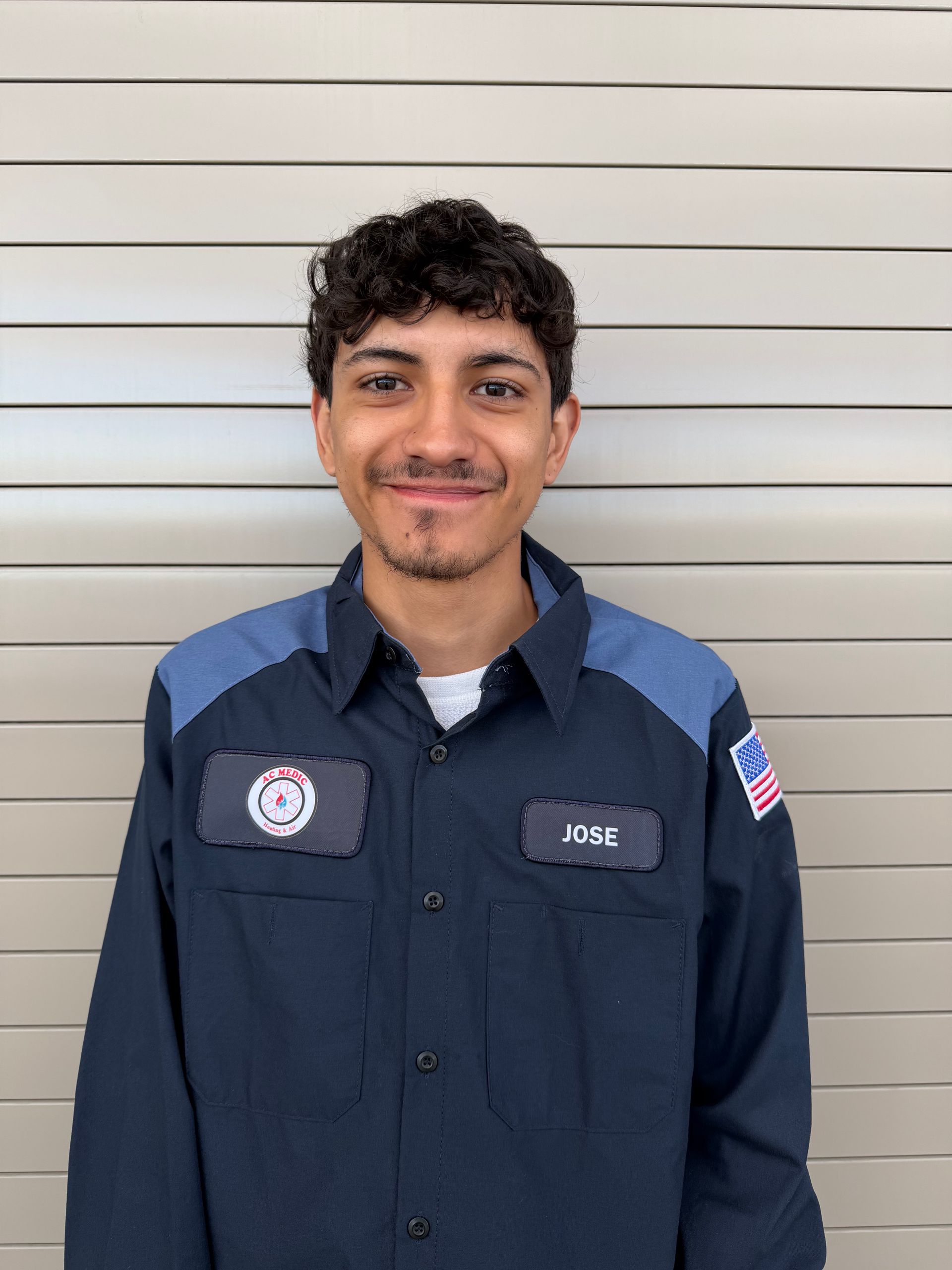 Man in work uniform smiles, posing in front of a light-colored, slatted wall.