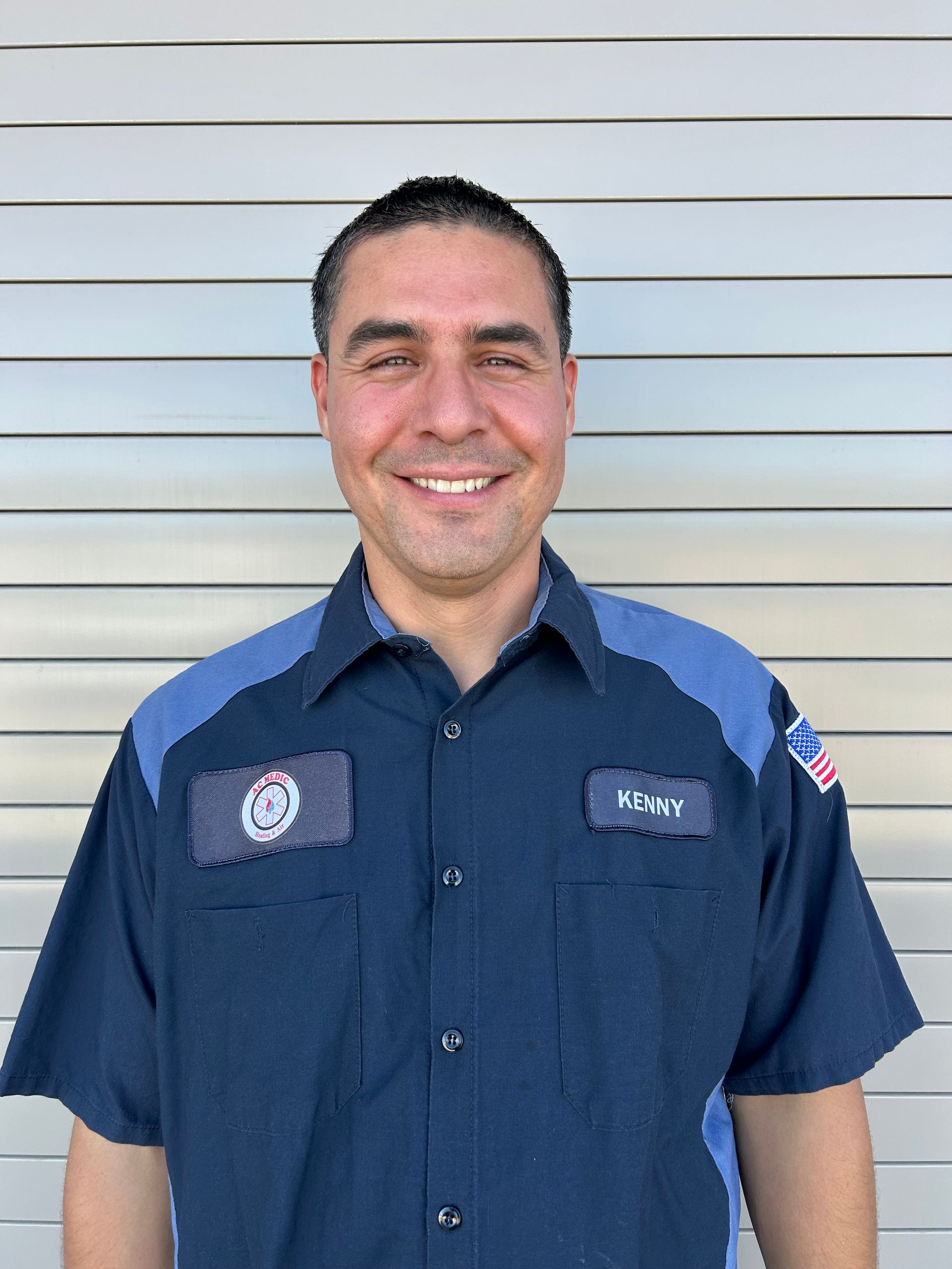 Man in blue work shirt smiling in front of a gray shutter background.