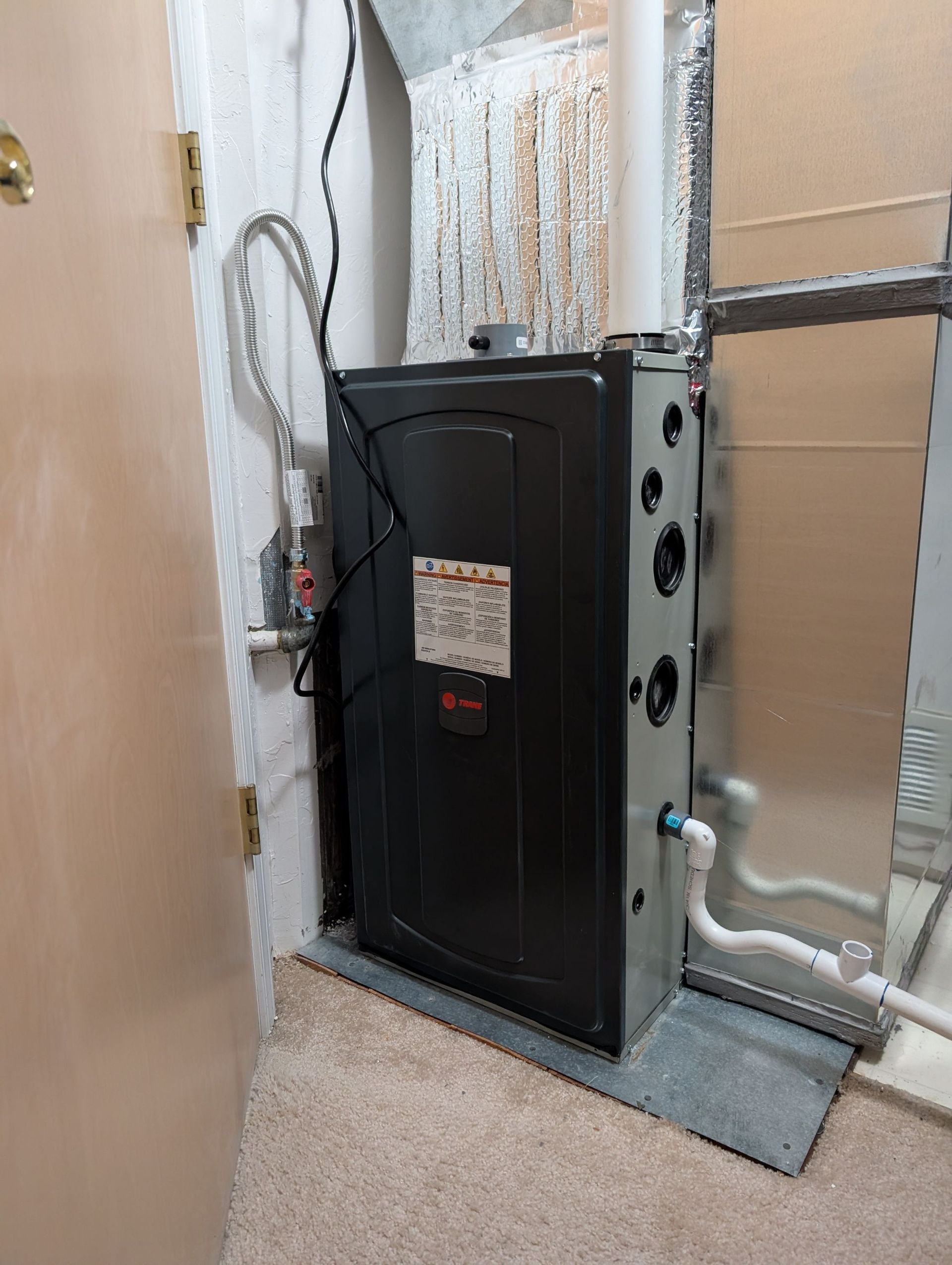 Furnace in a utility room. Black and gray appliance with pipes, ducts, and electrical components.