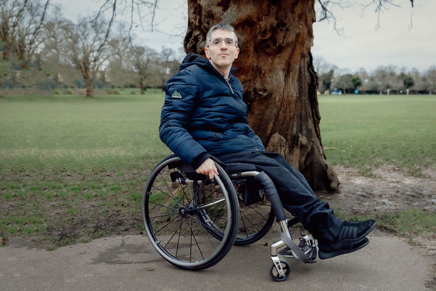 Founder of Dave Knows Disability David Bourroughs sitting in his wheelchair in a park by a true.  He is a white disabled man with glasses and in this photo is wearing a navy blue Superdry jacket.  He has his has on his wheels.