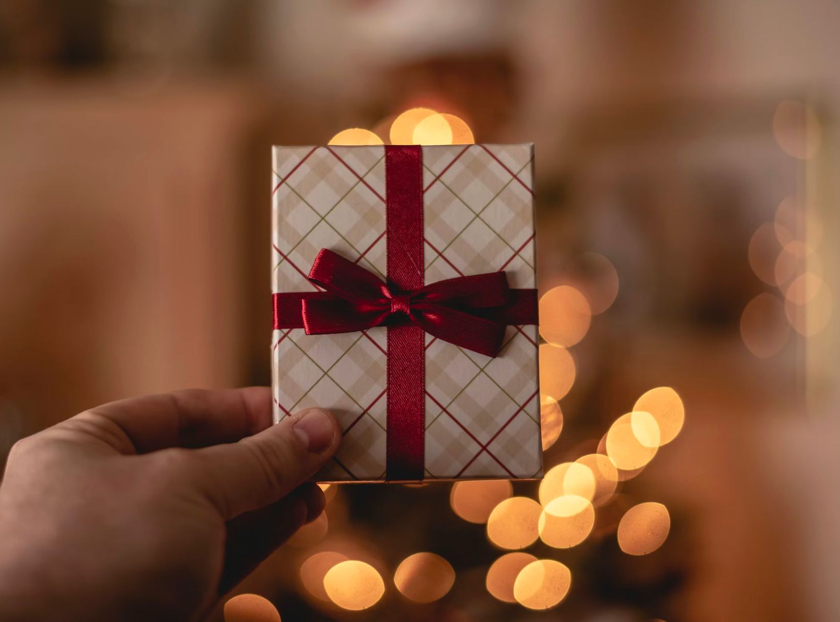 Hand holding a wrapped gift with a red ribbon, bokeh lights in the background.