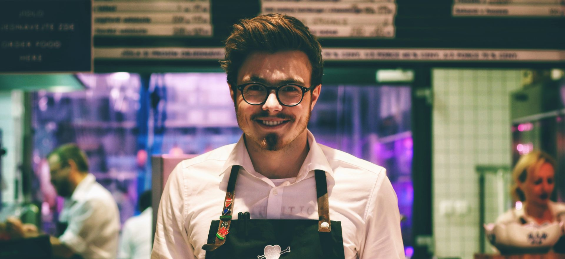Man wearing glasses and apron smiling in a cafe.