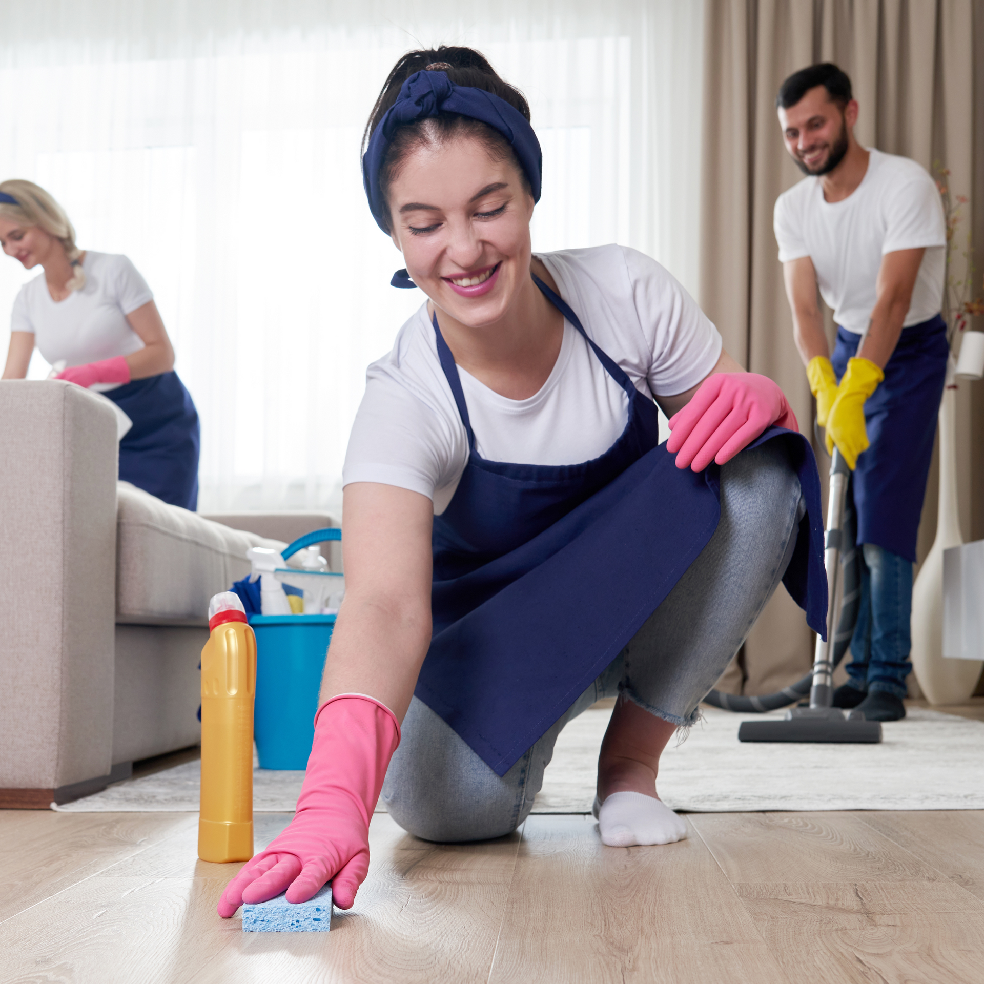 Three people cleaning a room; one wipes floor, other vacuums and the last cleans a couch.