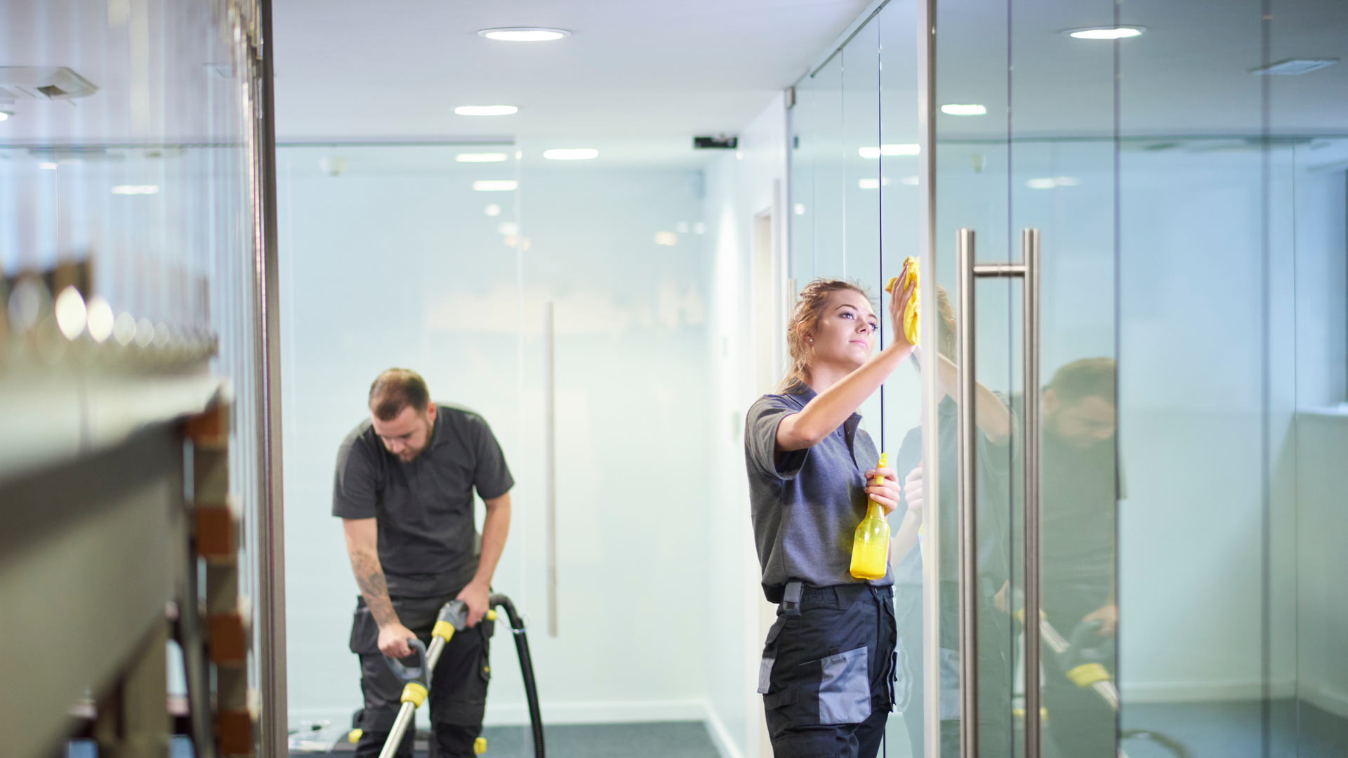 Two people cleaning an office: one vacuums, the other wipes a glass door. Both wear grey uniforms.