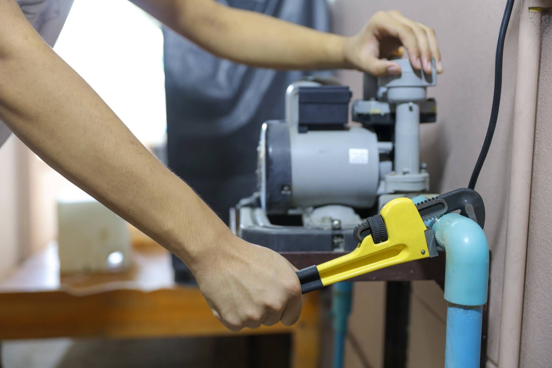 A person uses a yellow pipe wrench to tighten a connection on a gray water pump with blue pipes.