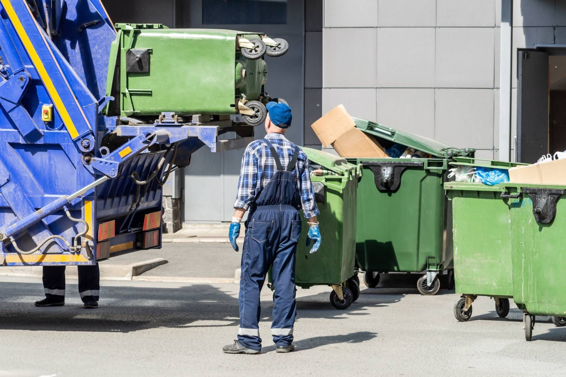 A worker in blue overalls stands next to a garbage truck as it lifts a green bin, with several other dumpsters nearby.