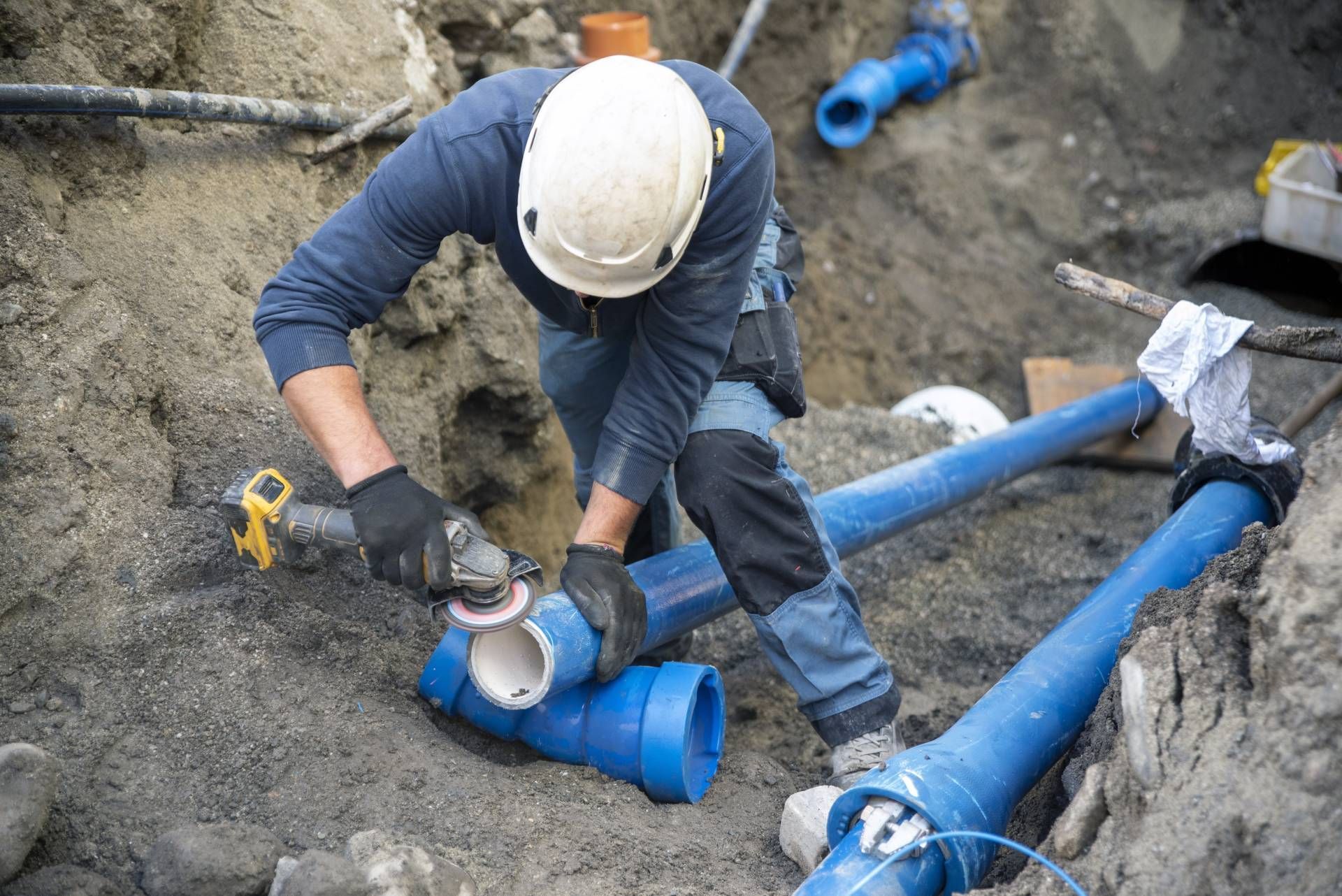 A construction worker in a white hard hat uses a power tool to cut a blue pipe in a dirt trench.