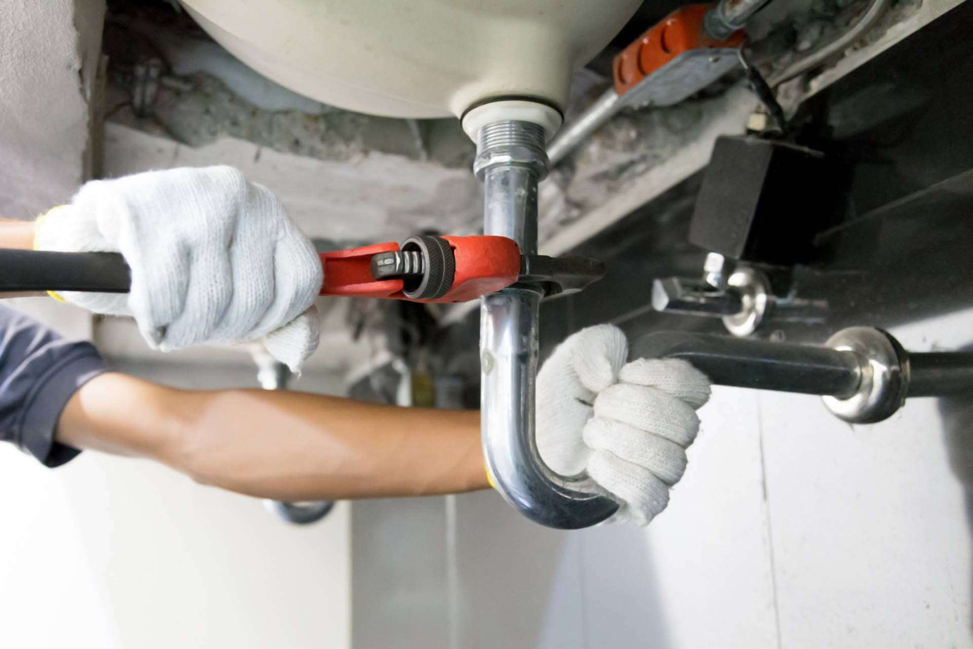 A plumber in white work gloves uses a red pipe wrench to tighten a metal pipe under a sink.