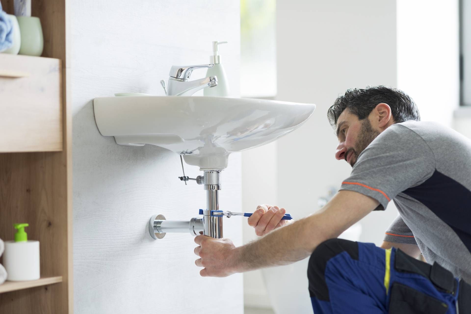 A plumber in uniform uses a wrench to repair the P-trap pipe underneath a wall-mounted bathroom sink.