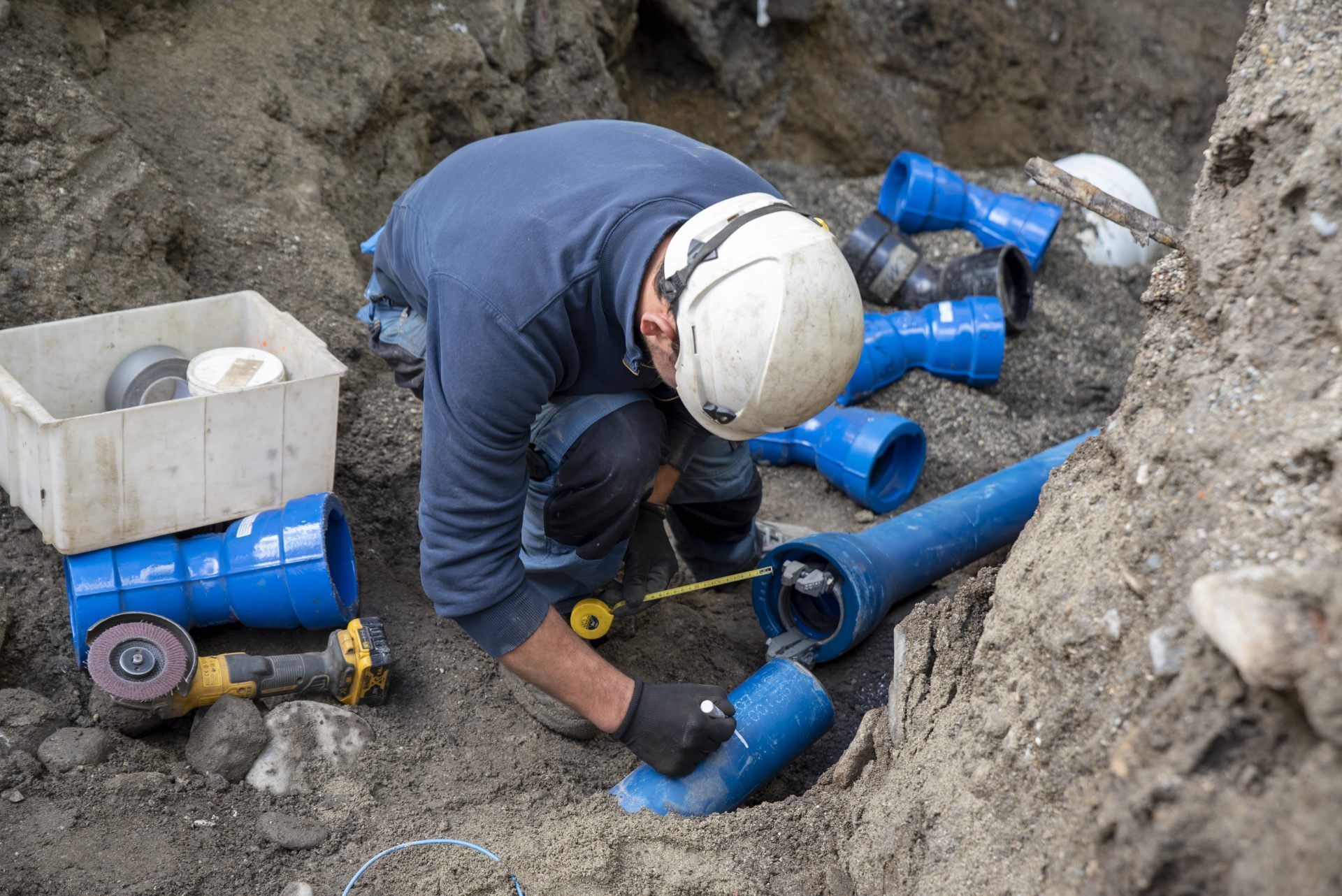 A construction worker in a hard hat measures a blue pipe inside an excavation trench filled with various pipe fittings.