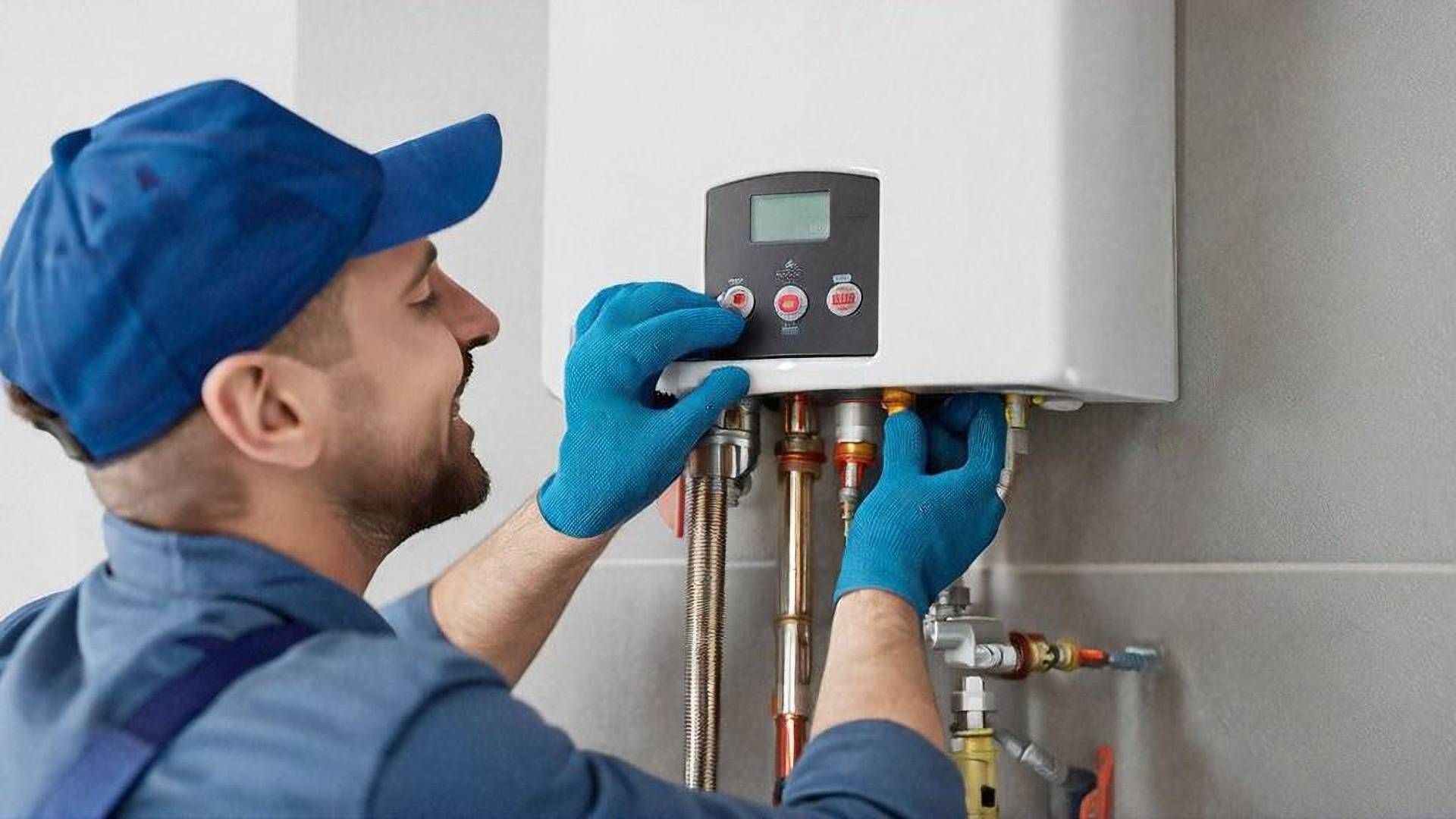 A service technician in a blue uniform and gloves adjusting the digital control panel on a wall-mounted boiler.