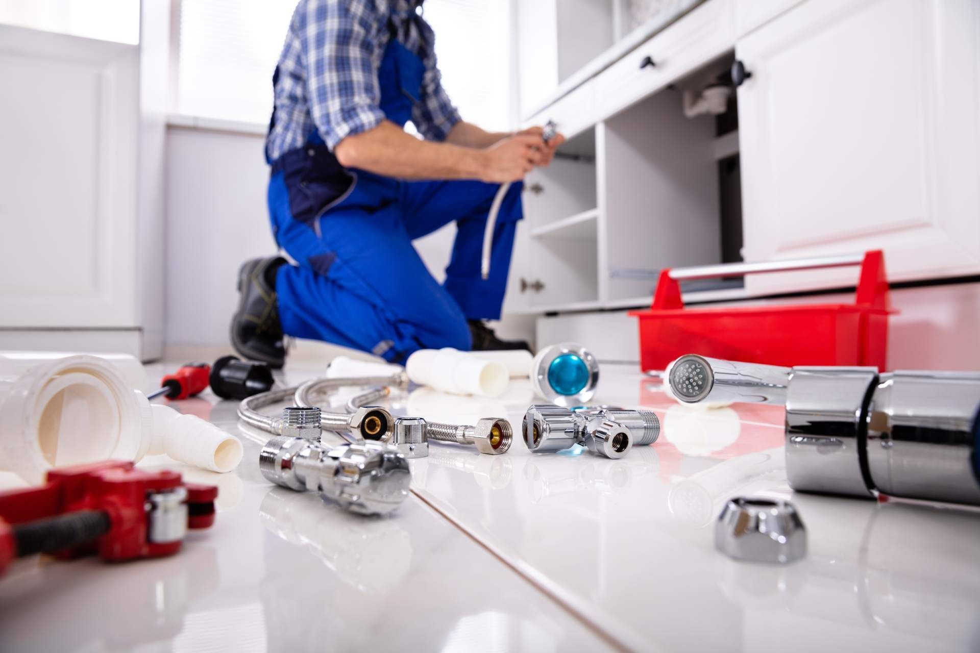 A plumber in blue workwear kneeling to repair kitchen cabinet plumbing, with various tools and parts spread on the floor.