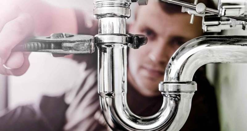 A focused person uses an adjustable wrench to tighten a metallic P-trap pipe under a sink.