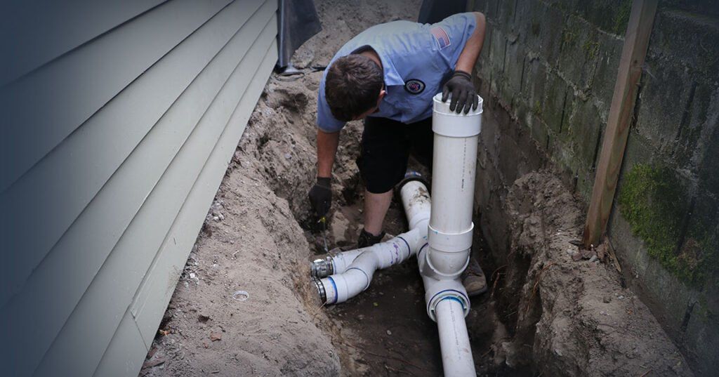 A person in work clothes connects PVC drainage pipes in a narrow trench between a house wall and a brick fence.