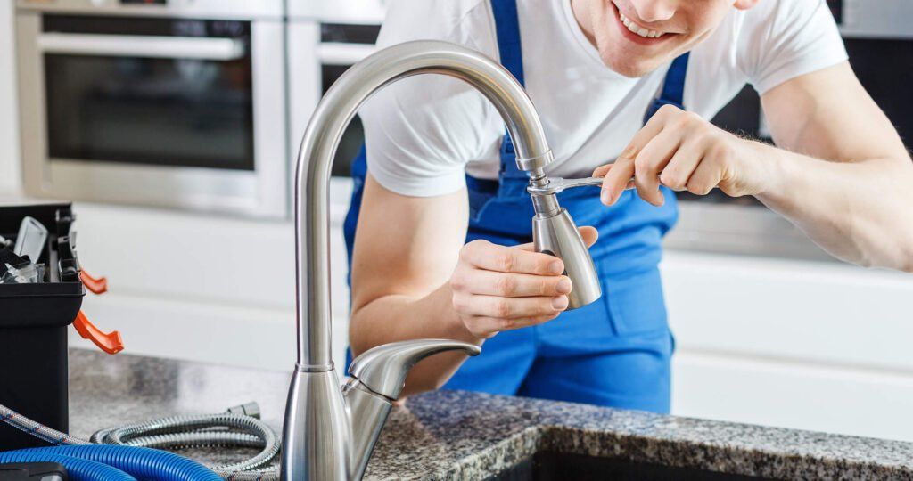 A smiling plumber in blue overalls repairing a brushed-nickel kitchen faucet at a granite countertop.