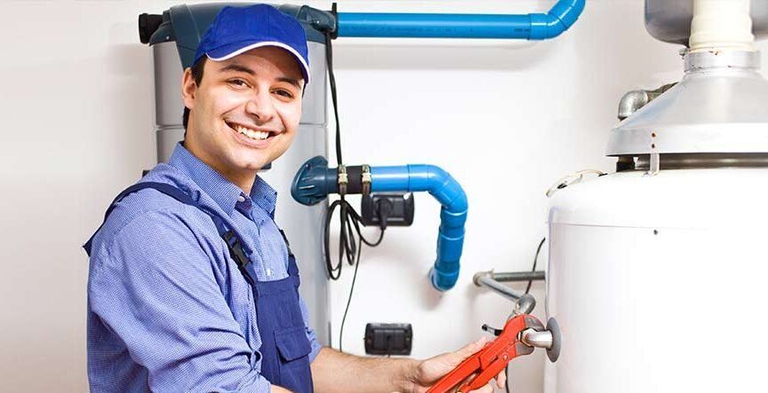 A smiling technician in a blue uniform holds a red pipe wrench while working on a residential water heater.